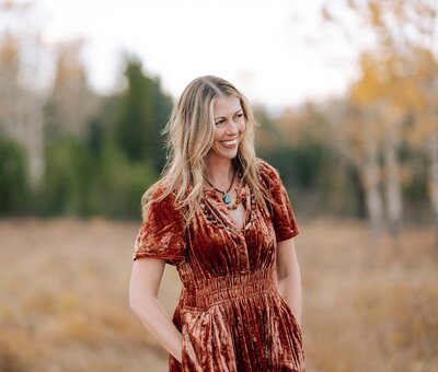Stevie looking away and smiling. Hands in the pockets of a auburn colored velvet dress. 
