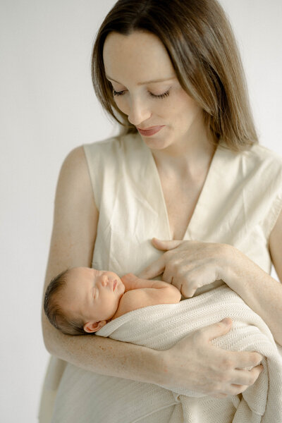 Studio newborn portrait of mom and baby wrapped in cream swaddle on minimal backdrop.
