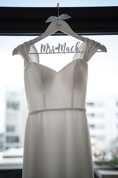 Elegant white wedding gown hanging on a custom “Mrs. Mack” hanger, backlit by natural light in a suite at JPS Events in downtown Indianapolis.
