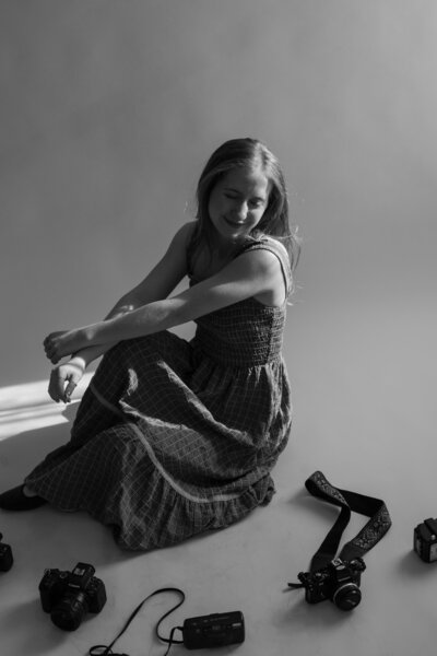 Autumn the wedding photographer sitting on a backdrop with cameras on the floor around her, in black and white.