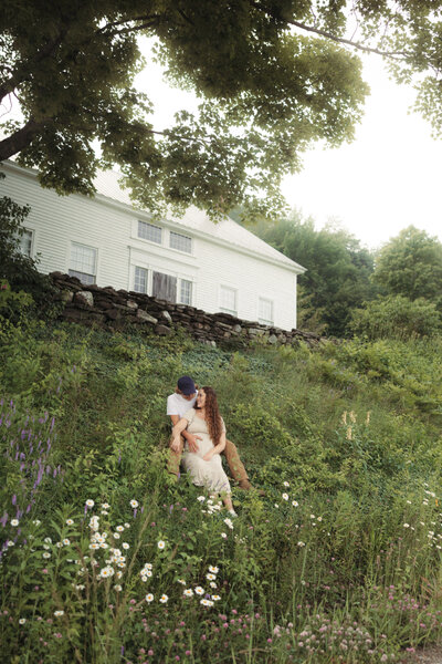 husband and wife sit together on grassy hill surrounded by wildflowers and a Vermont farm house in the background. Husband holds pregnant wife's belly and the two smile at eachother