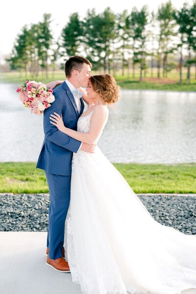 Bride and groom sharing a kiss by a lake, with the bride holding a bouquet of pink and white flowers, capturing a romantic moment.