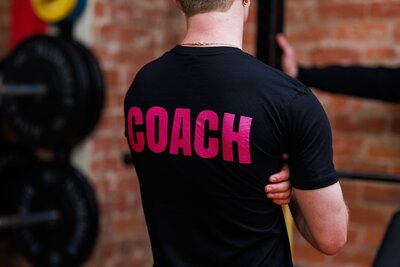 Coach supervising a pt session in a 24 hours dog friendly gym in Melbourne. Has the word Coach on his t shirt on the back.