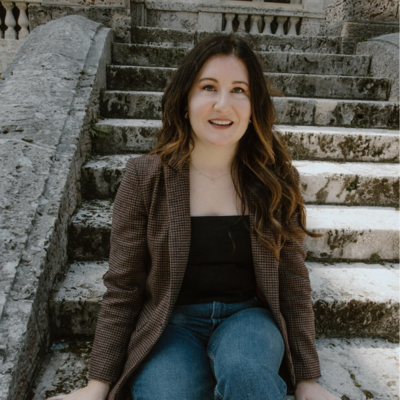 A woman with wavy brown hair sits on stone steps. She is wearing a plaid blazer, a black top, and jeans. She is smiling softly, and the steps and building behind her are made of stone, giving the setting a historical feel.