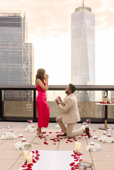 Romantic rooftop proposal in New York City with a view of One World Trade Center, as the groom kneels surrounded by rose petals and candles, captured by NYC wedding photographer Heritage House.