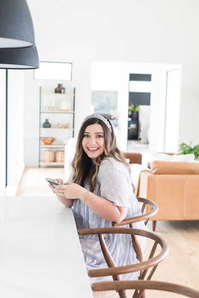 Real Estate Advisor is sitting  on a modern wooden chair. Behind her is the living room with a beautiful decoration and light orange couch. she has her phone and is wearing a dress and bandana.