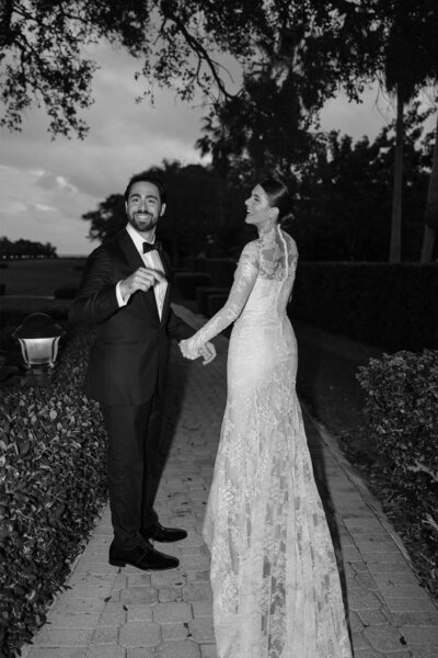 Bride and groom holding hands and smiling as they walk down a tree-lined path at dusk.