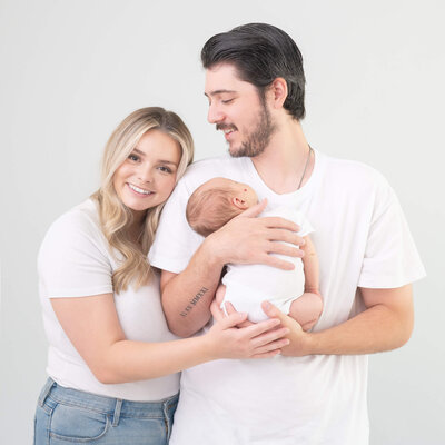 beautiful mom and dad in white with newborn in white against white backdrop captured by Allison Amores Photography