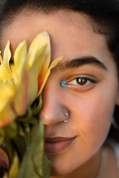 A close-up portrait of a young woman partially hidden behind vibrant yellow flowers, showcasing her piercing gaze, nose jewelry, and a pop of blue eyeshadow. The warm golden light highlights her natural beauty and expressive features, creating a striking and artistic senior portrait by Megan Bowers Photography.