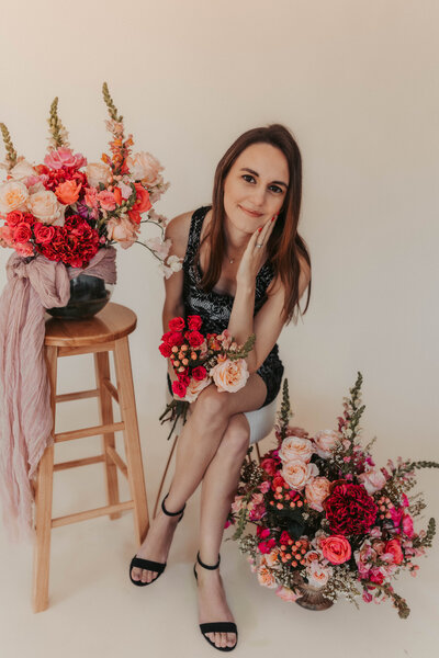 Woman wearing black dress posing sitting on stool leaning forward with face on hand and surrounded by pink and white flowers