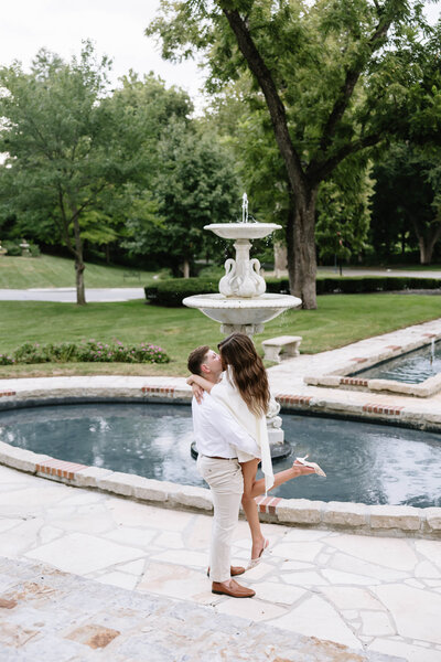 A couple kisses passionately under an ornate lace umbrella. The woman rests her hand on the man's face, conveying romance and intimacy in a lush park setting.