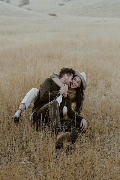 Couple sitting and laughing together in a yellow grass field during their sunset engagement session.