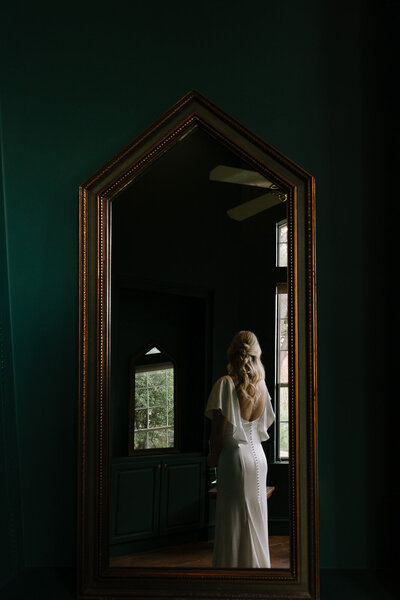 Bride looks into tall vintage gold mirror just before the wedding ceremony.