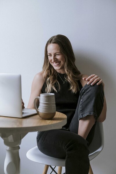 Woman sitting at her laptop, laughing and smiling, with a cup beside her, enjoying a moment of relaxation and engagement.