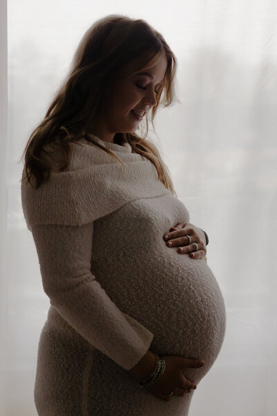 Een zwangere vrouw staat voor een raam, met een serene uitstraling en een hand op haar buik.