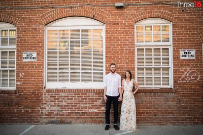 Engaged couple hold hands as they pose in front of a brick building in a alley in Old Towne Orange