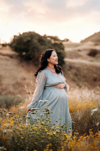 mom in gold maternity dress looks down at belly in a field of wildflowers