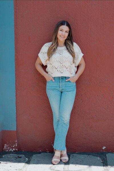 Natalie standing against a terracotta red wall in a cream knit top and jeans, smiling with hands in pockets, ready to connect.