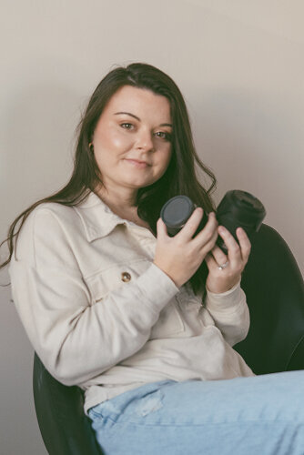 Michaela of Michaela Crawford Photography is facing the camera and smiling softly. She is holding her camera and a lens in her hands.