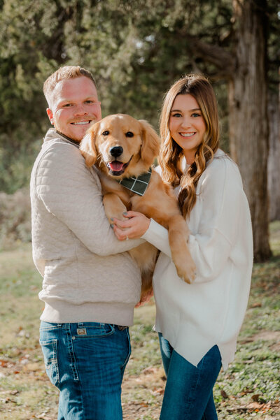 A couple is captured by the camera holding their dog and smiling.