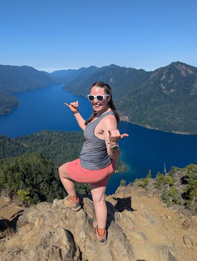 Sage Trails Alexandria Headshot - photographer sharing hang loose signs at the top of a mountain over looking a lake in Washington State.