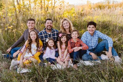 Family sitting in a sunlit meadow with fall colors, everyone smiling and relaxed during a warm outdoor portrait session.