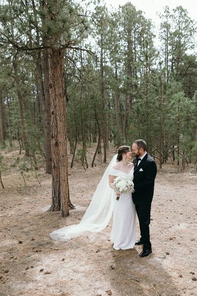 A bride and groom are standing amongst tall pine trees and looking at each other with smiles on their faces. 