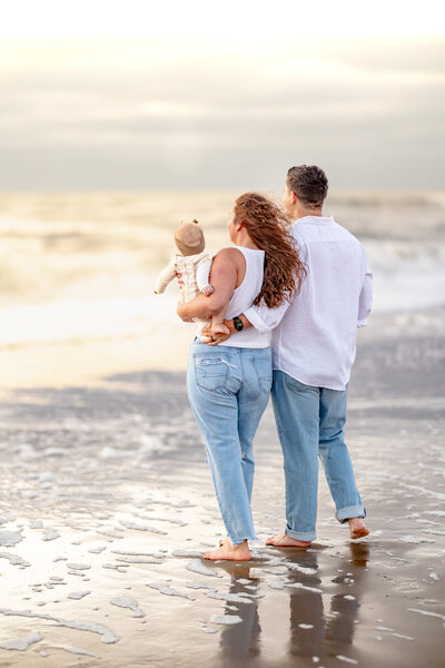 A mother and father hold their newborn close while standing in the shore break at sunrise during a tender family session by Brooke Tucker Photography in Virginia Beach