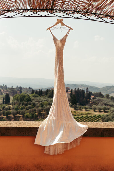 The wedding dress hangs gracefully, framed by the enchanting backdrop of Tuscany hills, blending the beauty of the gown with the serenity of the landscape