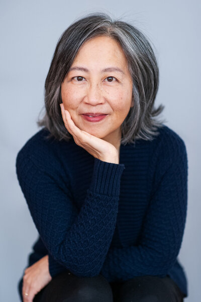 Studio headshot of an Asian female author leaning head in hand.