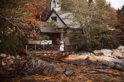 A bride and groom stand on a deck on top of a rocky coastline on a Maine island with their chapel where they got married in the background.