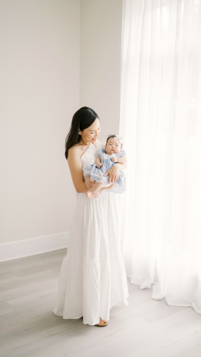 Mom in white dress holds baby girl in blue dress during photo session in Raleigh NC studio