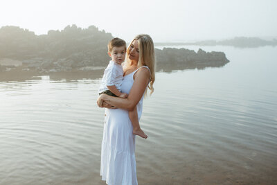 Family Phototgrapher on the gold Coast , a mothe ris holding her son on a beach at sunrise , the boy is turned looking at the camera and perched on his mother's hips. They are cuddled close with beach and water in the background