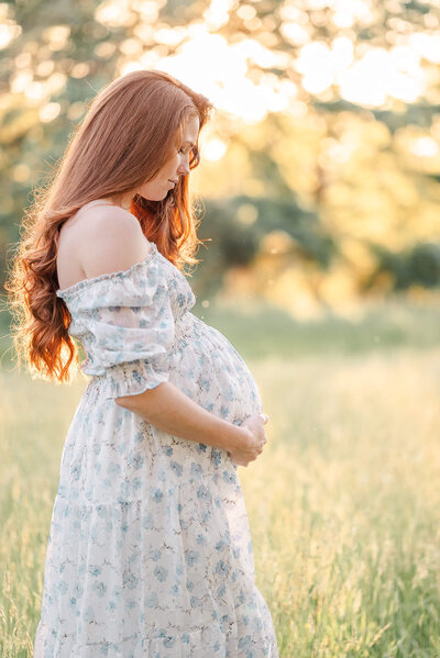 An expectant mother wearing a white floral dress, cradles her belly during her maternity session with Chesapeake Photographer, Justine Renee Photography.