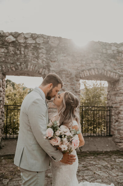 Bride and groom pose for wedding day photos at The Cambridge Mill in Cambridge, Ontario. The bride and room are standing in mill ruins with rich stonework and arches where windows used to be. The bride and groom's noses are touching and about to kiss. Captured by London, ON-based top wedding photographer and videographer Ashlee Ellison