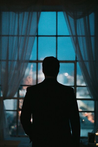 Silhouette of man in suit looking out large window at city lights