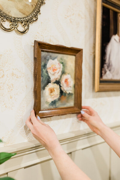 Female DIYer hanging a framed oil painting of flowers on the wall above board and batten trim.