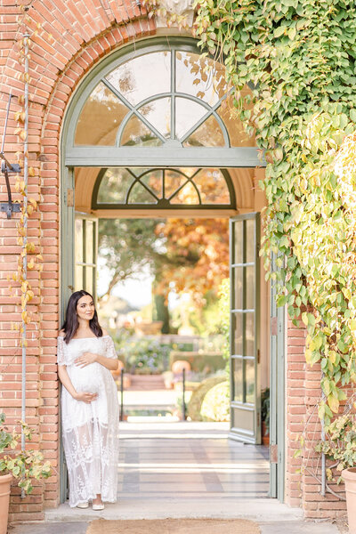 Pregnant woman in a long white dress holding her belly and leaning against a brick wall in a garden