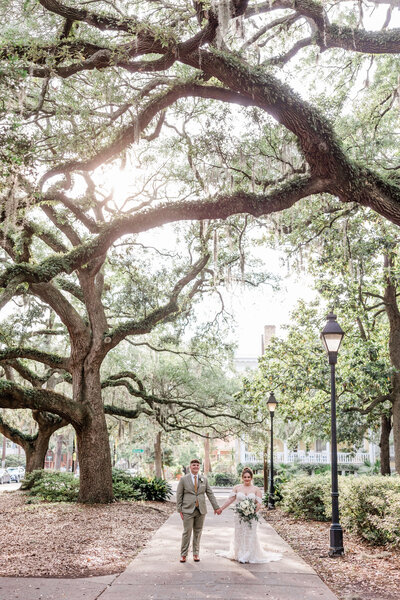 Bride and groom standing together under moss-draped oak trees in Savannah, GA, with the bride holding a neutral bouquet.