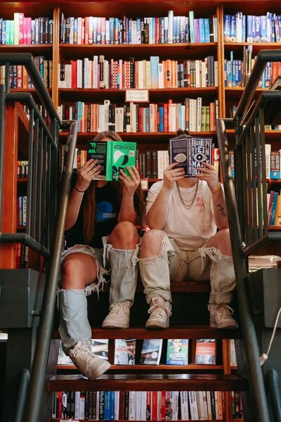KP and Jessie hold books up to cover their faces in a bookstore.