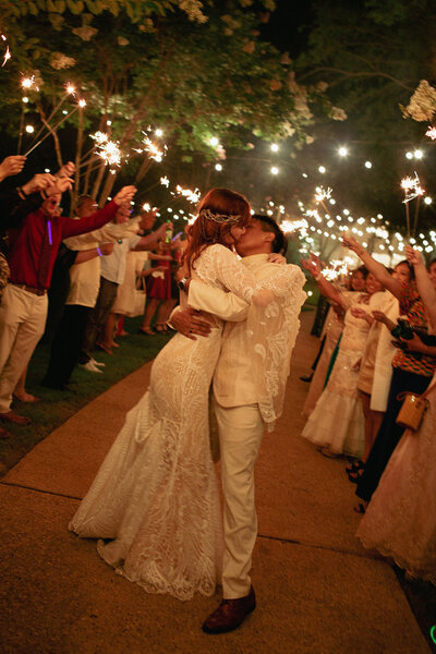 A couple shares a kiss in the middle of a path while guests hold sparklers overhead. The bride wears a lace gown, and the groom is in a light-colored suit. The scene is festive and illuminated by the warm glow of the sparklers.