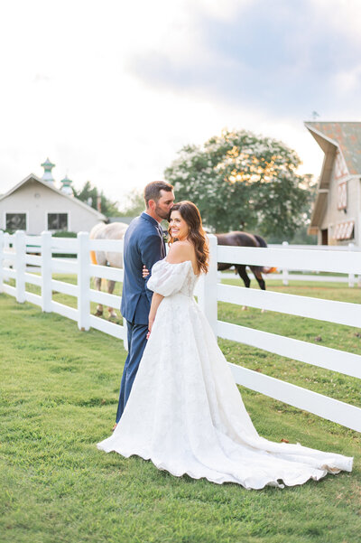A groom who is kissing the head of his bride. The bride is looking back towards the camera and similing. They are standing in front of a white farm fence with horses behind them.