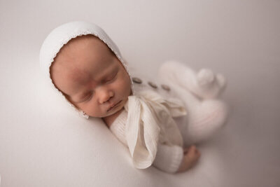 Close-up of a newborn baby sleeping peacefully, dressed in a white knit outfit and bonnet, lying on a soft white blanket.