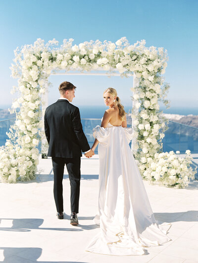 bride and groom in front of white rose ceremony arch overlooking santorini, santorini wedding photographer, greece wedding photographer, europe wedding photographer, destination wedding photographer