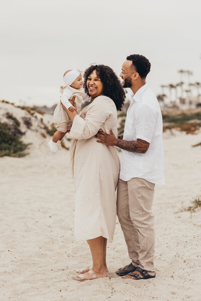 mom holds up her adorable baby girl while dad smiles at them on the beach in Coronado