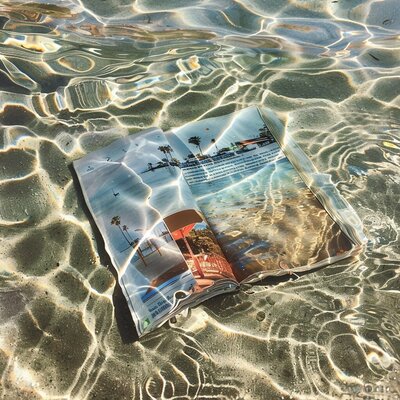 An open magazine resting underwater in a shallow, sandy area, with sunlight creating shimmering patterns across its colorful pages depicting vacation destinations. This artistic shot emphasizes the inspiration and wanderlust sparked by vacation fundraising for charities, offering a creative way to support impactful missions.