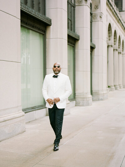 black groom in white tux jacket walks among stunning architecture during wedding photos in Detroit, Michigan
