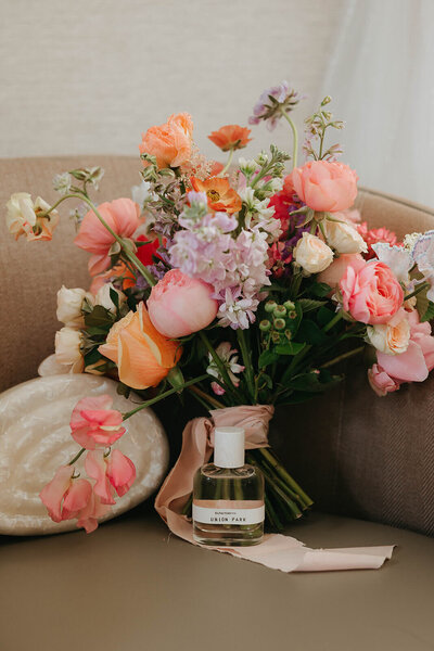 Garden-style bouquet in coral, peach, and pink tones resting on a velvet chair beside a ribbon-tied bottle of perfume and a shell dish.
