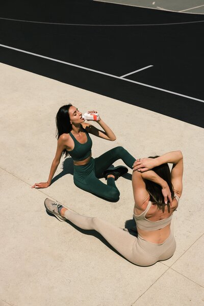 two woman sitting on the floor of a tennis court drinking a beverage.the floor of a tennis court