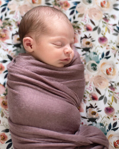 Newborn girl in a purple swaddle laying in her crib with floral sheets.